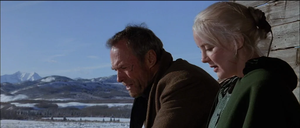 Unforgiven (1992) movie still: medium — A snowy mountain landscape serves as the backdrop to a wood structure. A man and a woman sit together…