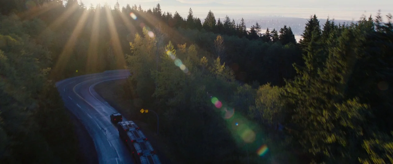 Deadpool 2 (2018) movie still: wide — A dark mountainous highway curves through a dense evergreen forest during the golden hour. A convoy of dark…