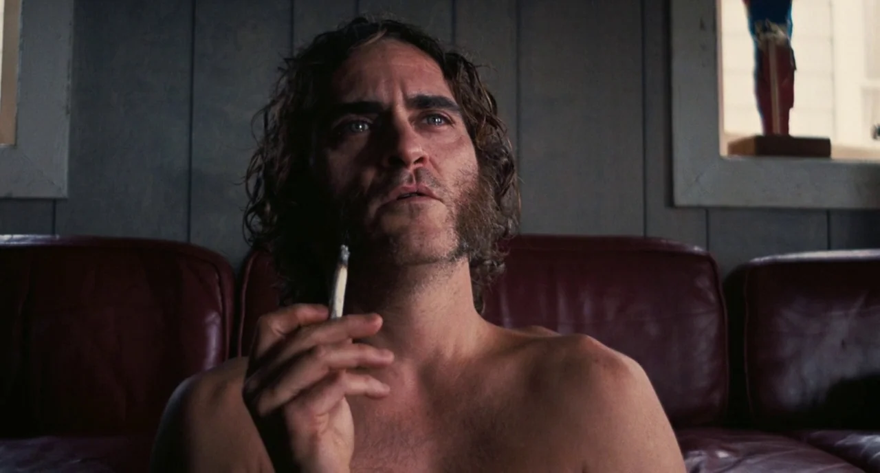 Inherent Vice (2014) movie still: medium — An interior space features wood-paneled walls and a worn, dark red leather couch. A shirtless man sits on…