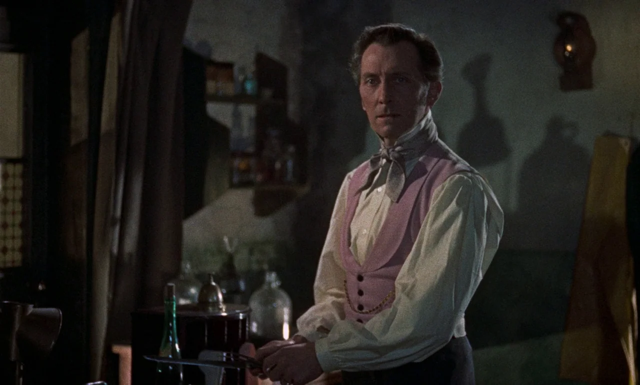 The Revenge of Frankenstein movie still: medium — A dimly lit room contains various glass vials and equipment in the background. A man stands in the…