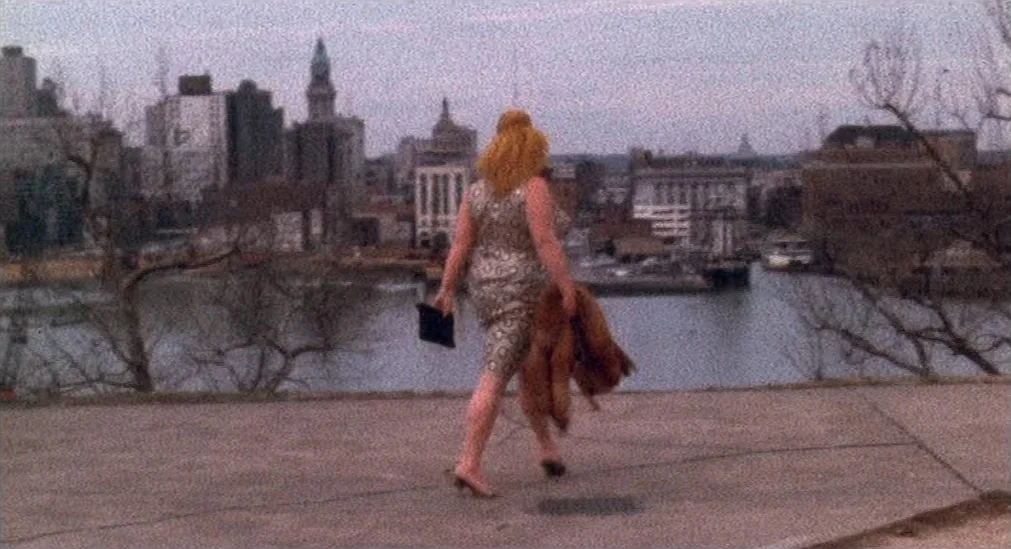 Pink Flamingos (1972) movie still: fullbody — A concrete embankment overlooks a wide river with a cityscape in the background. A woman in a…