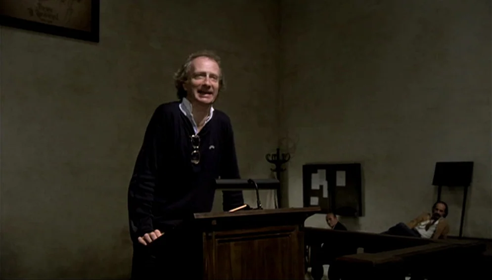 Orchestra Rehearsal (1978) movie still: medium — A dark, sparsely furnished courtroom with plain beige walls and wooden pews. One man speaks from a…