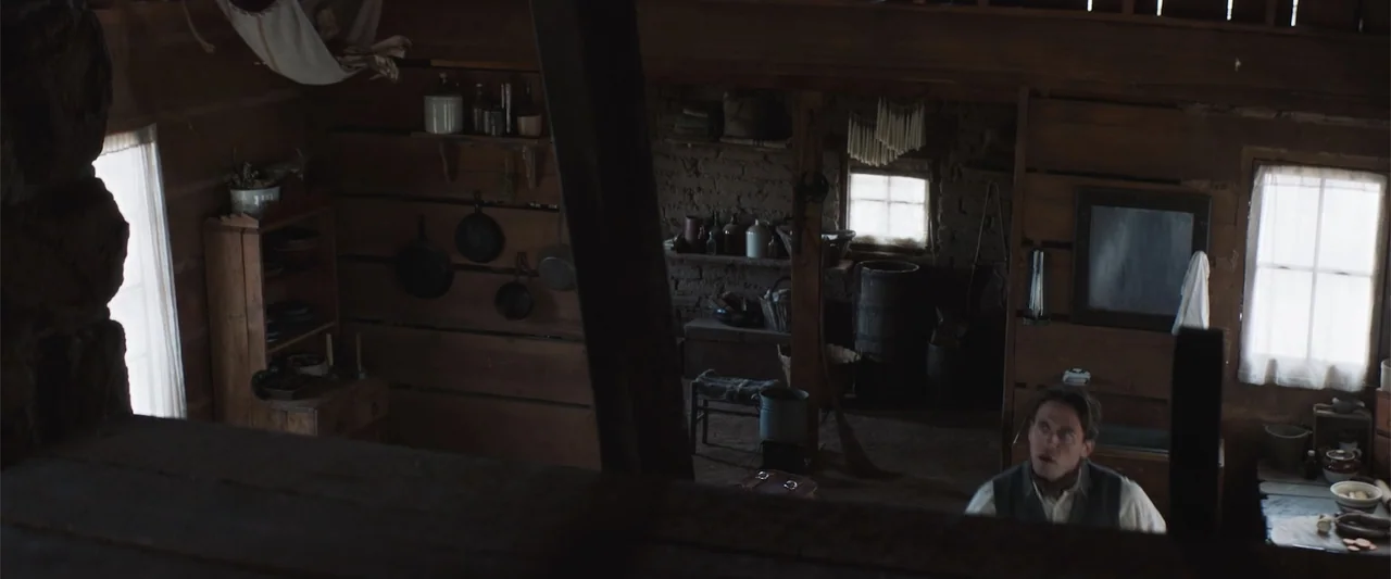 The Wind (2005) movie still: wide — A rustic log cabin interior with mud walls, iron cookware, and wooden beams visible. A man looks upward toward…