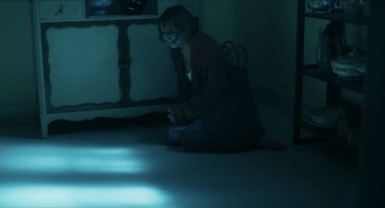Echoes Of A Crime (2025) movie still: medium — A dark, dimly lit interior room featuring a wooden cabinet and open shelving. A woman kneels on the…