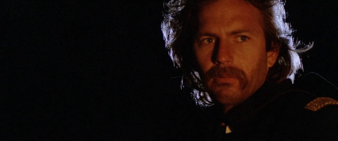 Dances With Wolves (1990) movie still: closeup — A dark night setting where a man stands against a pitch black background. The man gazes forward…