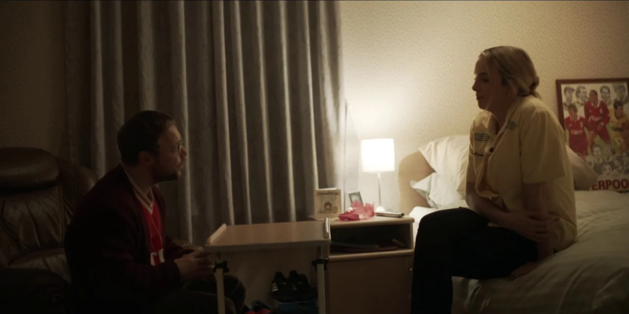 Help (2021) movie still: medium — A dimly lit room contains a bed, a bedside table, and heavy gray curtains. A woman in a yellow shirt sits on the…