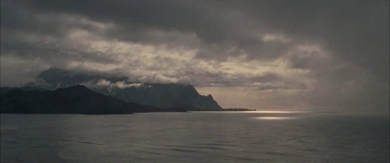 The Descendants (2011) movie still: establishing — A vast coastal view featuring dark, mountainous terrain under a heavy, overcast cloud bank. Light…