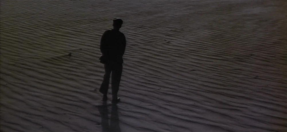 Lawrence Of Arabia movie still: wide — A vast sandy desert surface with distinct wind-rippled textures dominates the frame. A lone figure in…