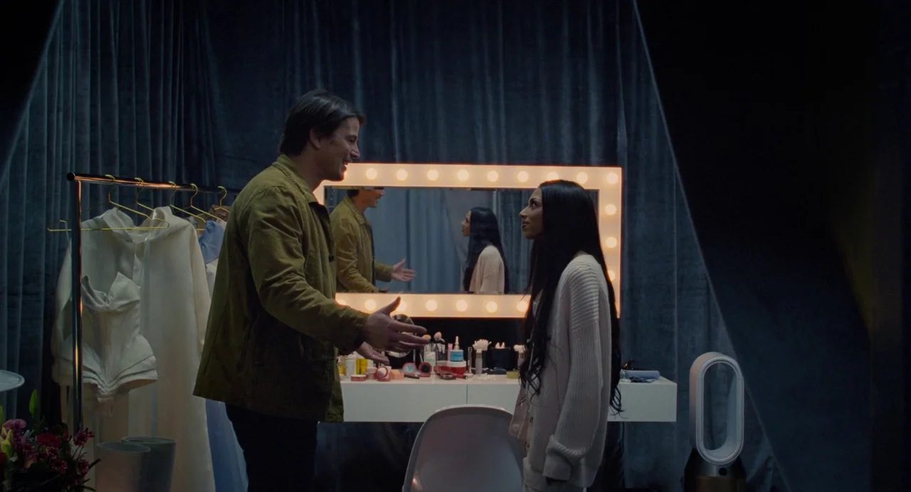 Trap (2024) movie still: medium — A dressing room features dark blue curtains and a large illuminated vanity mirror. A man stands gesturing towards…