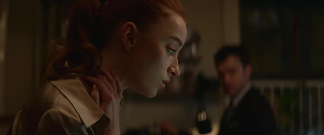 Fair Play (2023) movie still: closeup — A dimly lit room with an out-of-focus figure in the background. A woman in the foreground looks down at an…