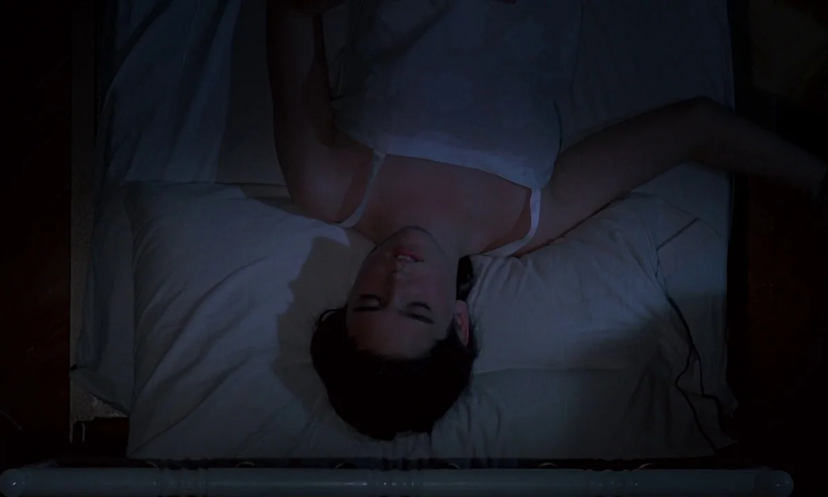 Phenomena (1985) movie still: medium — A person lies centered on a white bed sheet viewed from directly overhead. The dark surroundings fade into…
