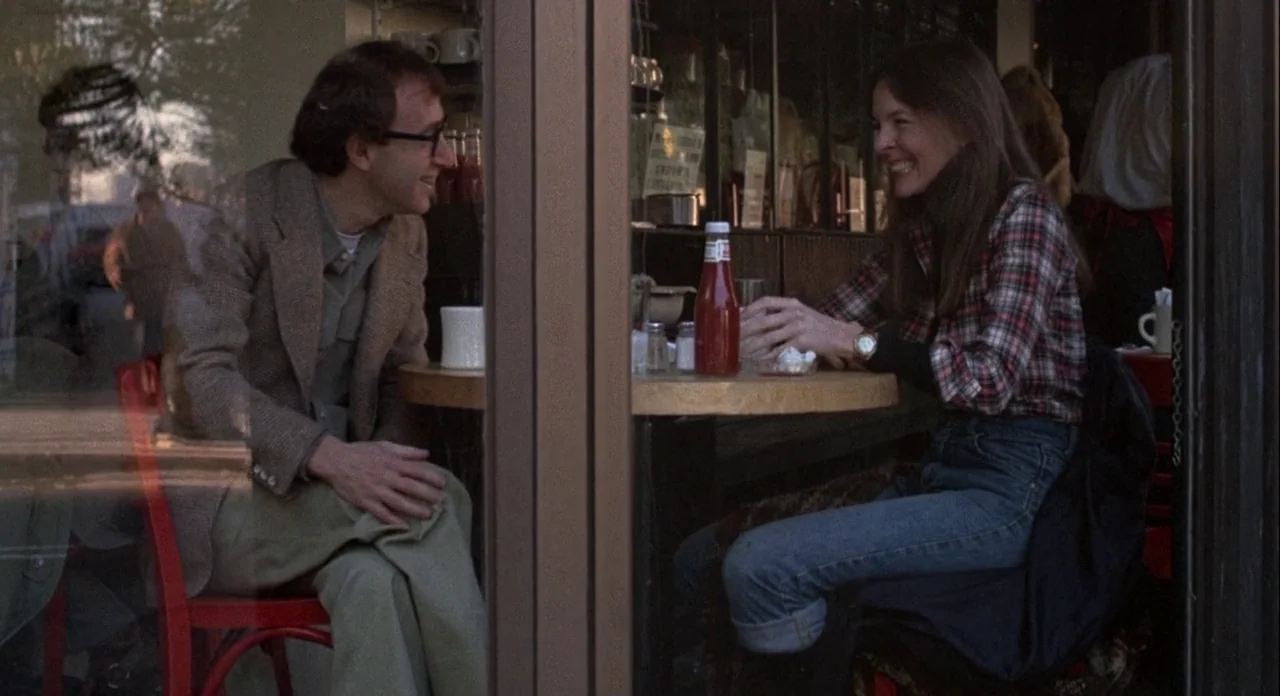 Annie Hall (1977) movie still: medium — A window view shows two people sitting at a wooden table in a restaurant. The man and woman face each other…