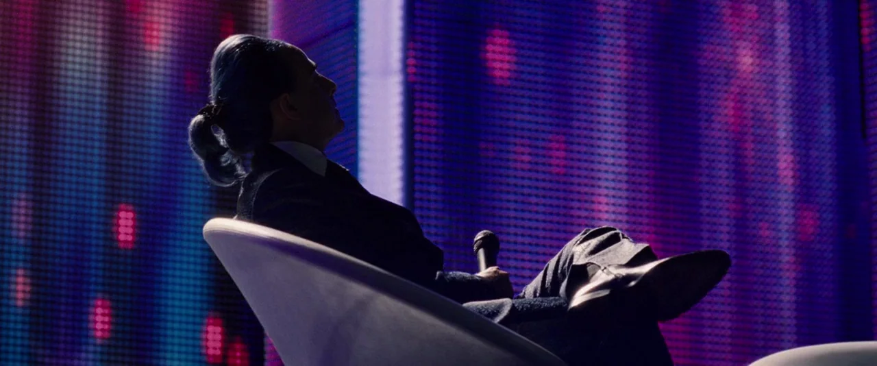 The Hunger Games (2012) movie still: medium — A television studio setting with a large pixelated LED backdrop glowing in violet hues. A man in a…