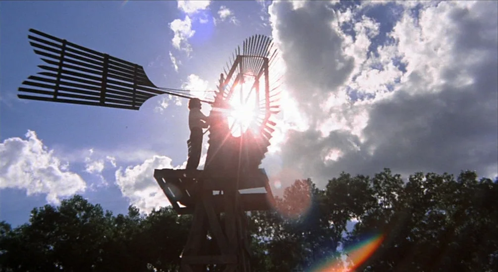 The Hired Hand (1971) movie still: wide — A tall wooden windmill stands against a bright sky with scattered white clouds. A person is standing on…