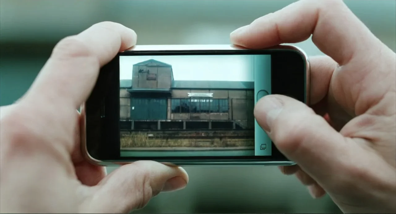 Soul Kitchen (2009) movie still: closeup — A person holds a smartphone displaying a photograph of an industrial building exterior. The device screen…