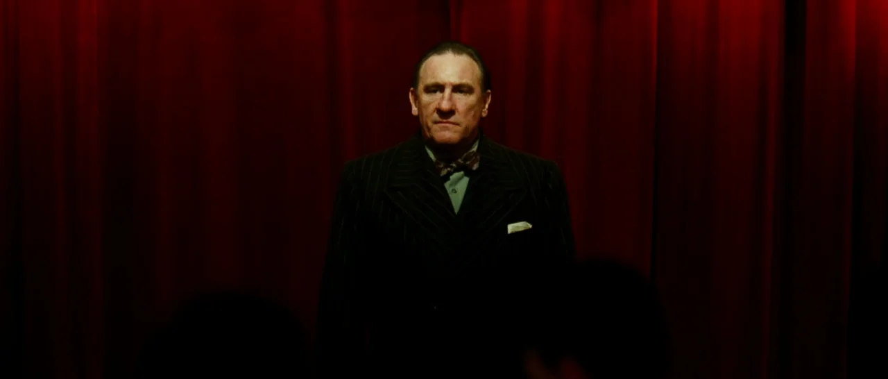 La Vie En Rose (2007) movie still: medium — A man stands centered before heavy red velvet curtains inside an indoor performance space. The subject…