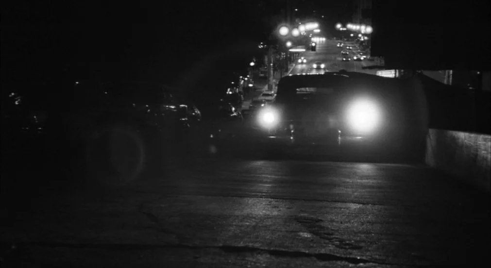Lenny (1974) movie still: wide — An asphalt road at night stretches toward distant streetlights and oncoming vehicle traffic. A vintage car drives…