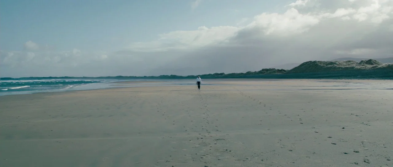 Calvary (2014) movie still: wide — A vast, empty tidal beach stretches toward a distant line of low dunes. A lone man walks across the wet sand…