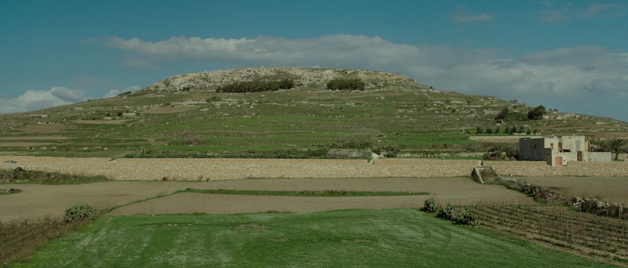 By The Sea (2015) movie still: establishing — A wide rural landscape features tiered green fields below a large limestone plateau hill. A small…