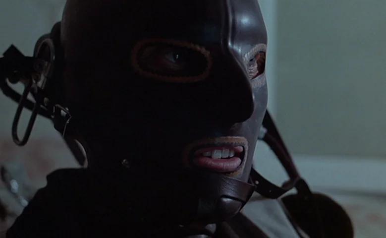 Maîtresse (1975) movie still: closeup — A dark leather mask with thick straps fits tightly around a human head. A person looks forward with…