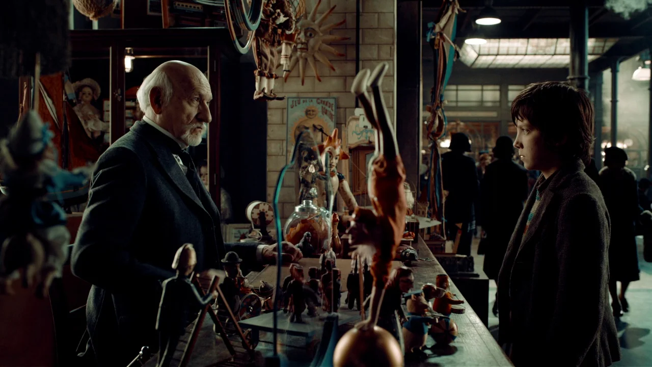 Hugo (2011) movie still: medium — A cluttered toy shop filled with vintage figurines and wooden playthings. An older man stands behind the counter…