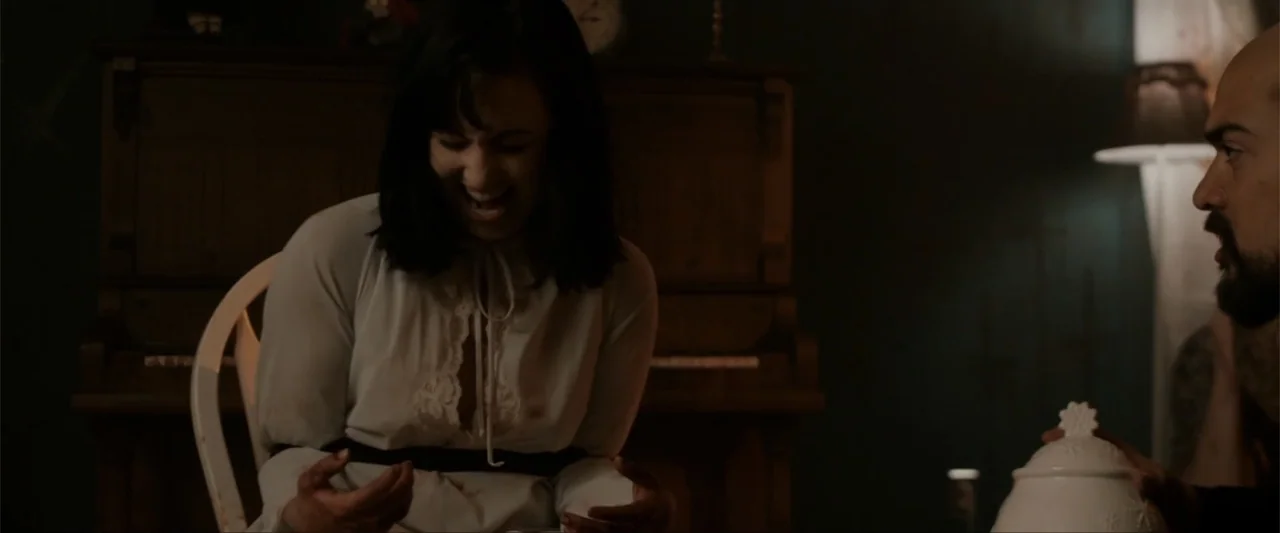 Anomaly (2006) movie still: medium — A dark room with a piano in the background and a table in foreground. A woman in a white blouse laughs while…