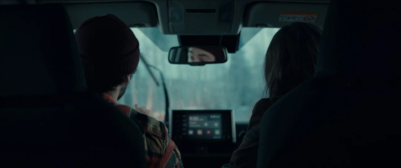 M3gan (2022) movie still: medium — The interior of a modern vehicle is viewed from the back seat looking forward. A man and a woman sit in the front…