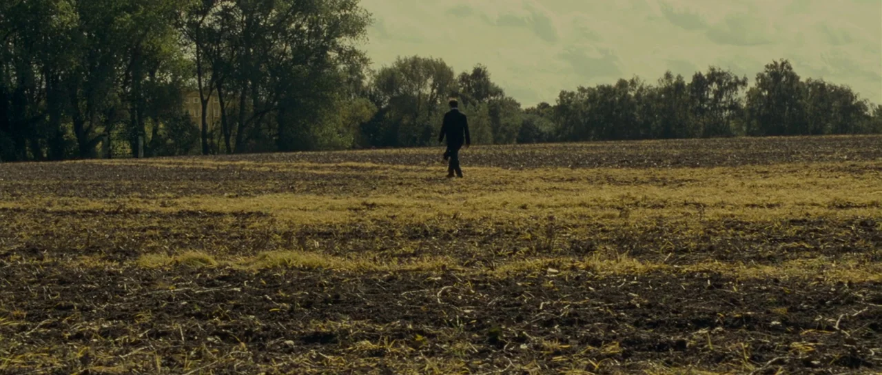 The Woman in Black (2012) movie still: wide — An expansive, harvested dirt field extends toward a dense line of trees. A lone man in a dark suit…
