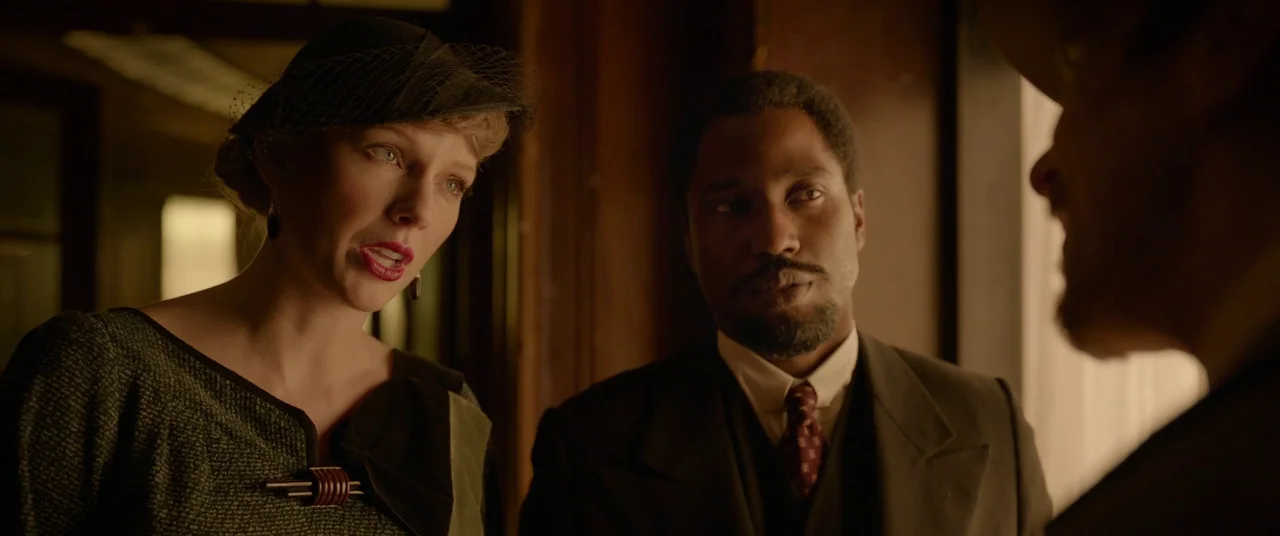 Amsterdam (2022) movie still: medium — A dimly lit room with dark wooden paneling serves as the backdrop for interaction. A woman in a 1940s hat and…