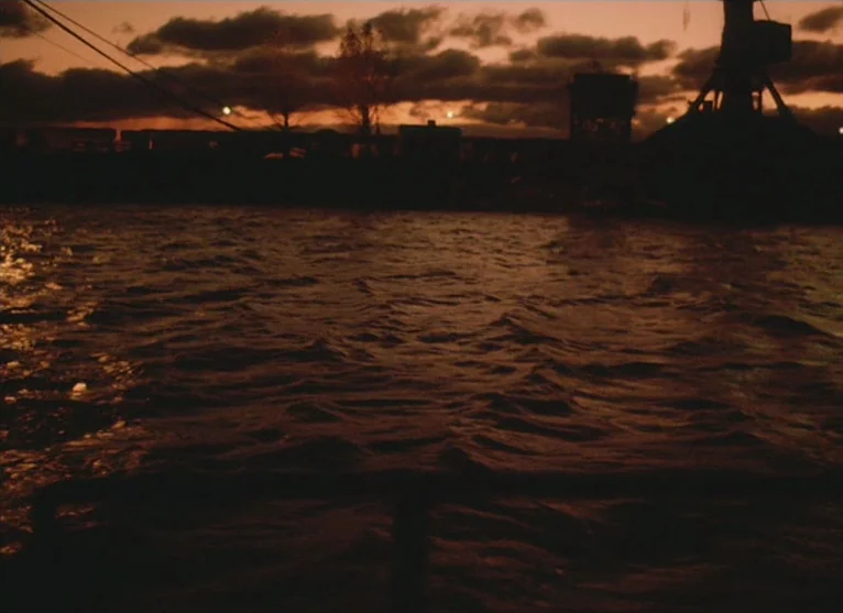 A Visitor to a Museum (1989) movie still: establishing — A dark harbor view at sunset features rippling water under a cloudy orange sky. Industrial…