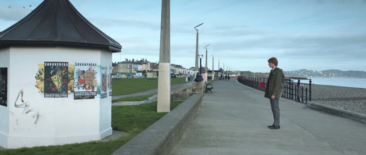 Frank (2014) movie still: wide — A concrete walkway borders a pebbled beach under an overcast, grey sky. A young man in a green jacket stands alone…