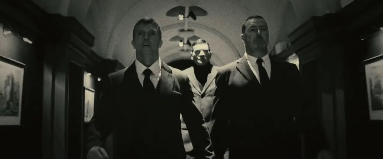 Trance (2013) movie still: fullbody — A dimly lit narrow vaulted hallway features framed art on dark paneled walls. Three men in formal attire walk…