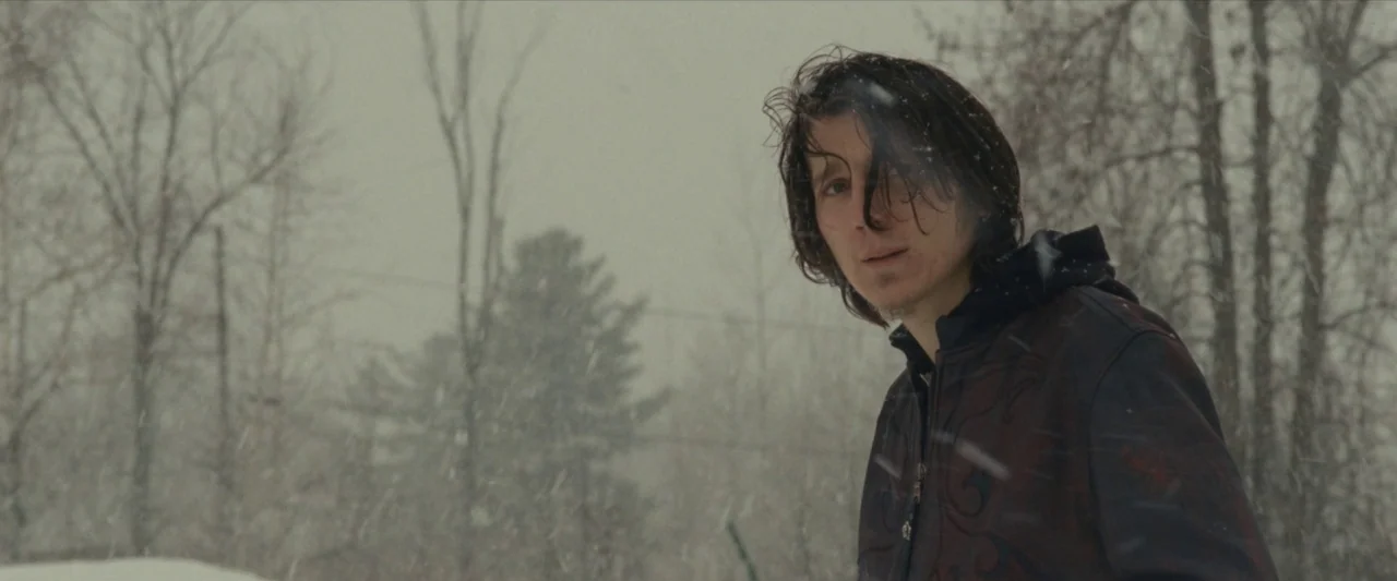 For Ellen (2012) movie still: medium — A snowy, overcast woodland environment with bare trees and falling snow. A young man stands in the…