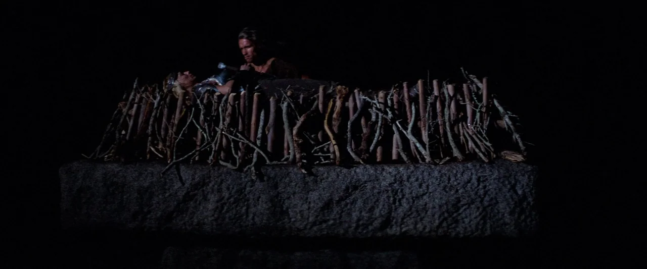 Conan The Barbarian (1982) movie still: wide — A raised stone platform is covered with a grid of dry wooden branches. A man kneels beside a woman in…