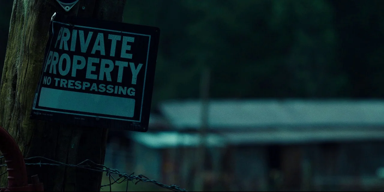 The Death of Dick Long (2019) movie still: closeup — A dark, blurred farm building sits in the distance behind a wooden utility pole. A black metal…