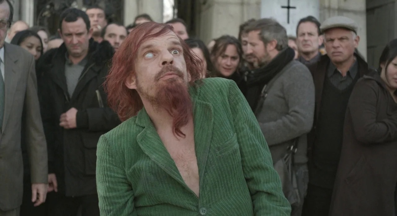 Holy Motors (2012) movie still: medium — A group of people stand behind a man in a green corduroy jacket outdoors. A stone tomb with a cross carved…