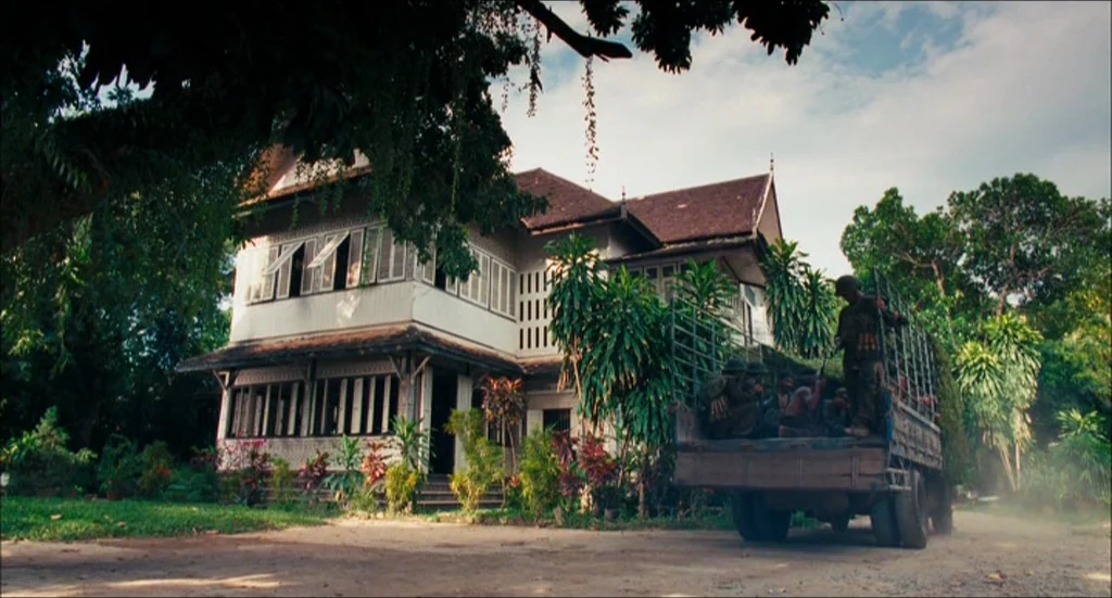 Rescue Dawn movie still: wide — A traditional two-story colonial mansion with white walls and dark shutters stands in greenery. A military truck…