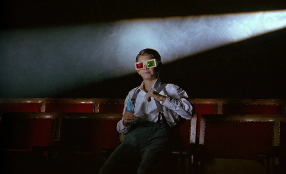Wittgenstein (1993) movie still: medium — A dark movie theater with empty rows of red velvet-backed seats. A young boy sits in the center wearing…