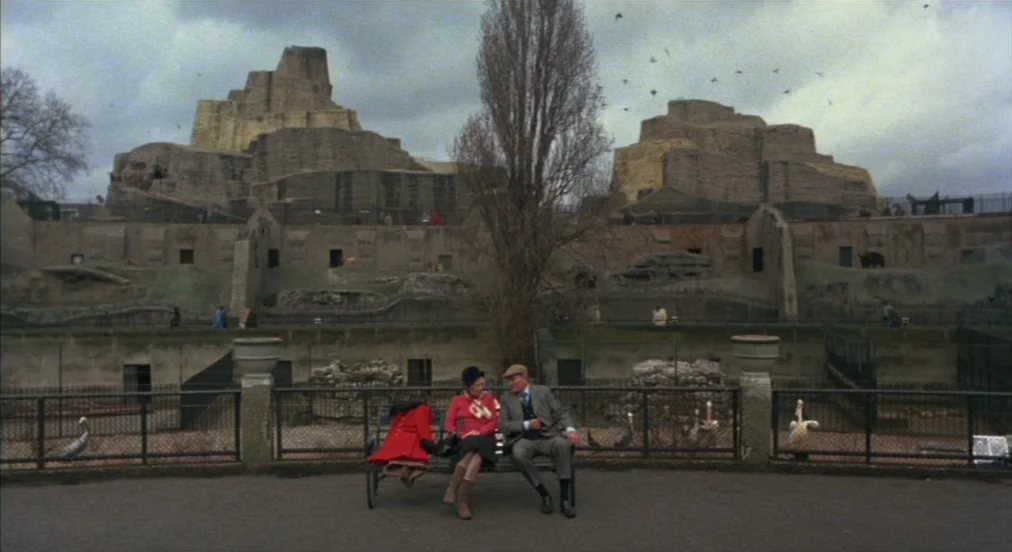 An American Werewolf in London (1981) movie still: wide — An outdoor zoo setting with large concrete animal enclosures and a cloudy sky. A man and a…