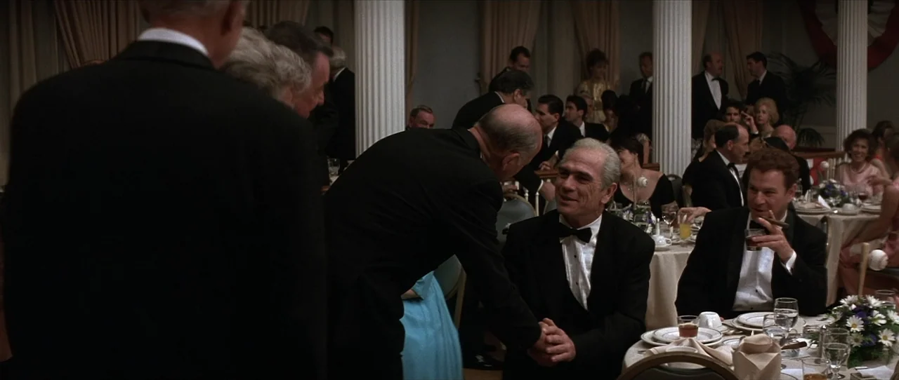 Cobb (1994) movie still: medium — A formal dinner hall features rows of round tables and white classical columns. A man in a tuxedo leans over to…