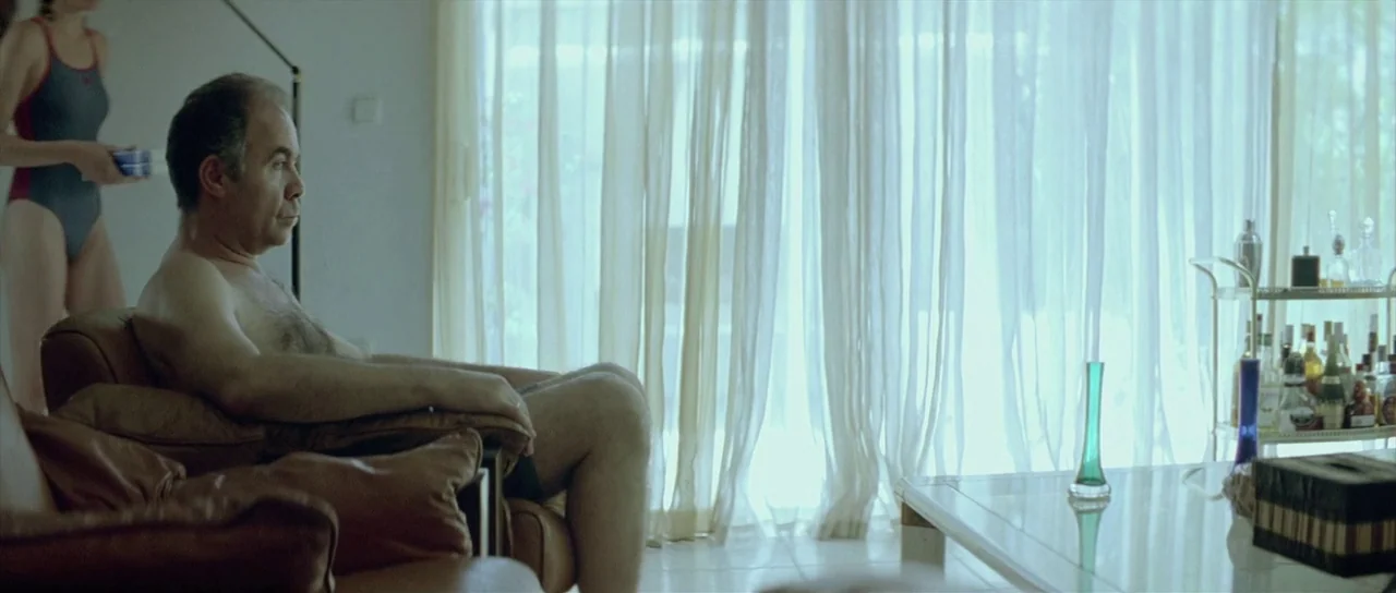 Dogtooth movie still: medium — A spacious room features sheer white curtains covering a large window wall. A middle-aged man sits shirtless in a…