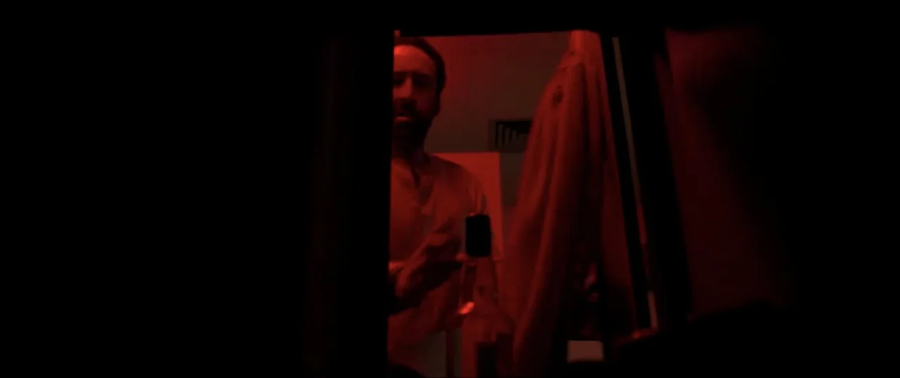 Primal (1994) movie still: medium — A dark bathroom interior is flooded with a monochromatic red light. A man stands in the doorway holding a glass…