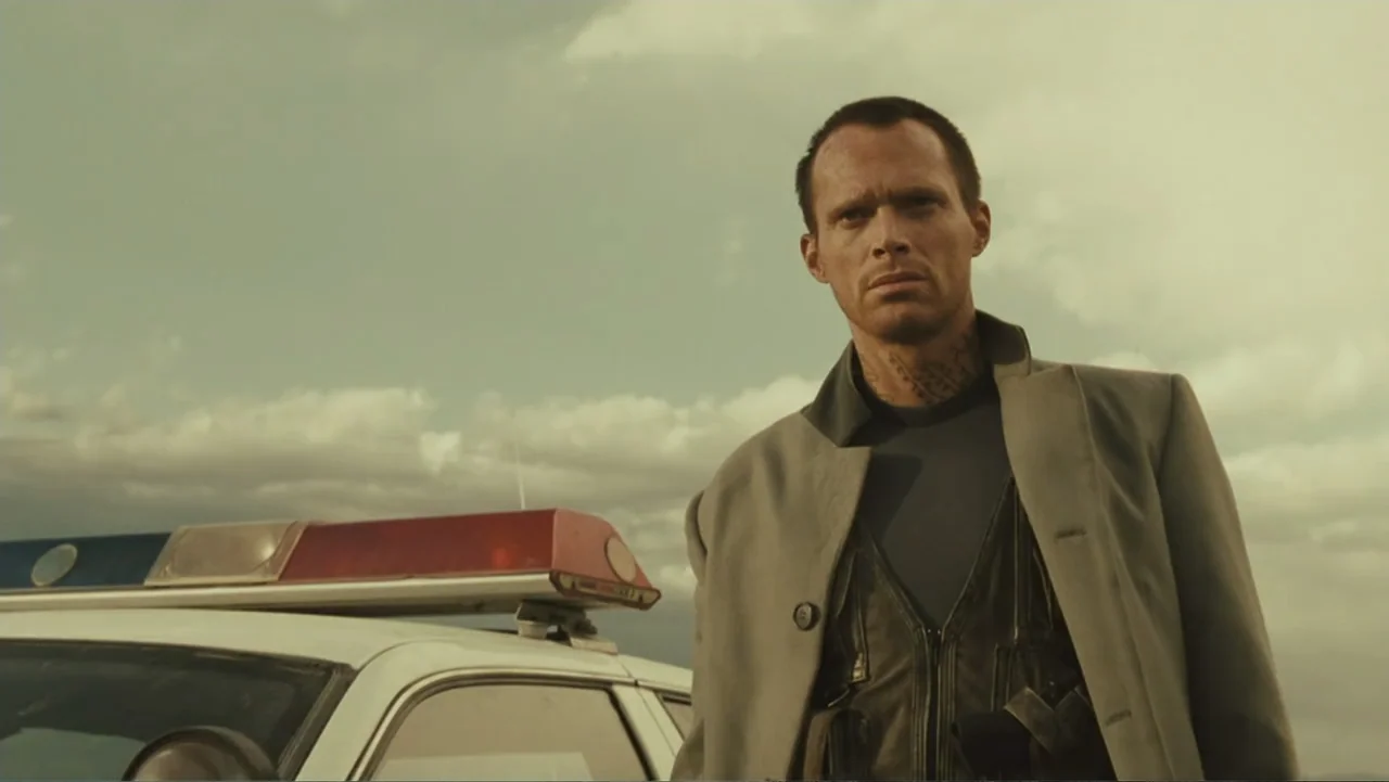 Legion (2010) movie still: medium — An open desert landscape with a police cruiser parked in the foreground. A man wearing a tactical vest stands…
