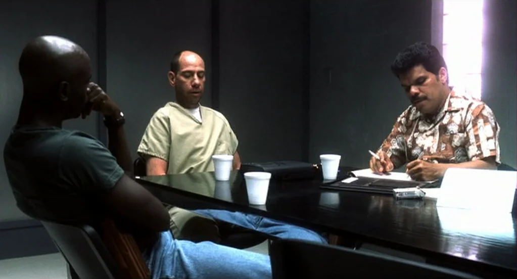 Traffic (2000) movie still: medium — An austere gray room with block windows and a dark table. Three men are seated, engaged in a professional…