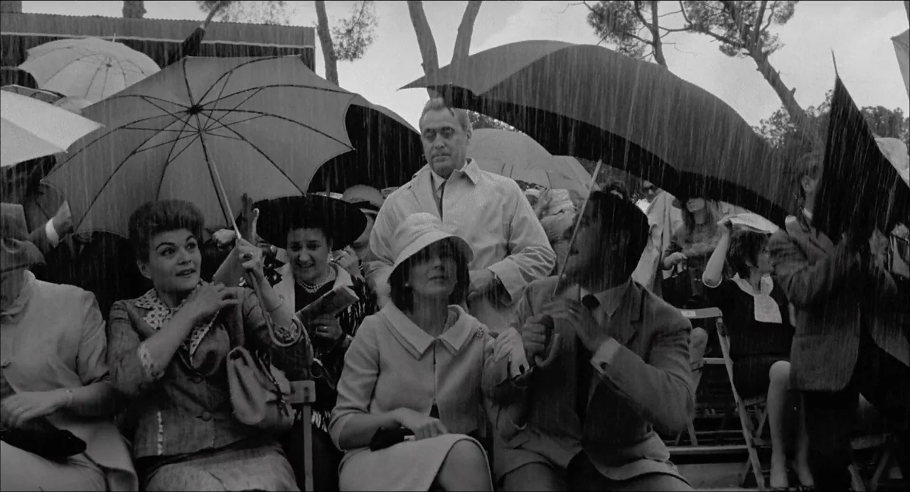 Il Boom (1963) movie still: medium — A group of people sit on metal stadium bleachers during a downpour of rain. Each person holds an umbrella to…