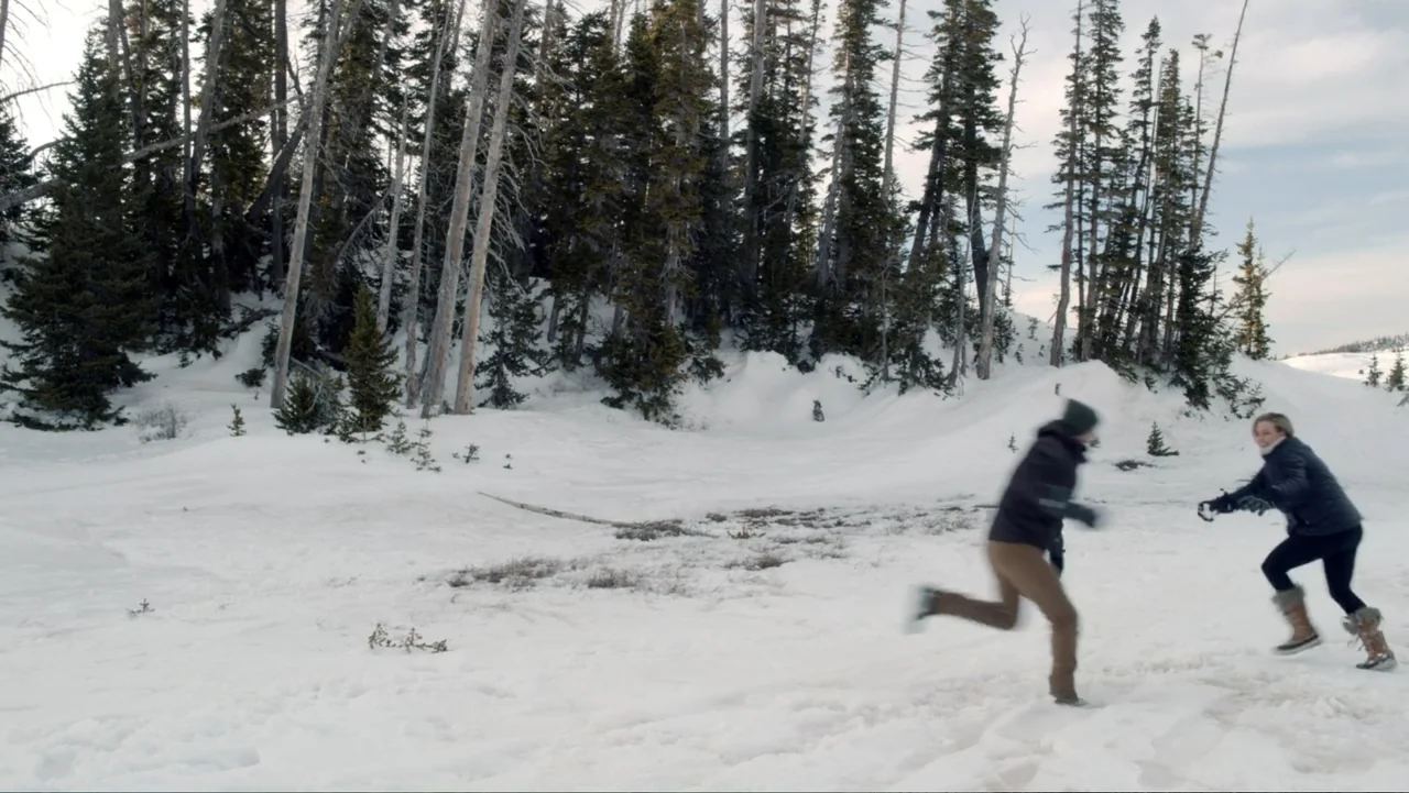 Alex & Emma (2003) movie still: wide — A snow-covered clearing sits in front of a dense pine forest with tall trees. A man and woman in winter gear…
