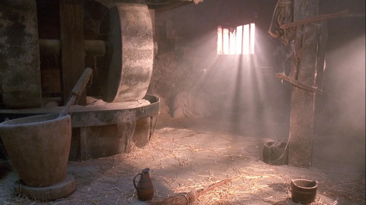 Army of Darkness (1992) movie still: establishing — An aged mill interior contains stone grinding wheels and scattered straw on the floor. Dust…