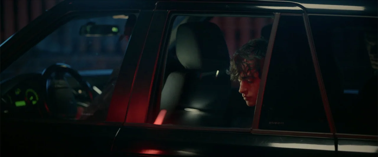 Love Me Love Me (2026) movie still: medium — The interior of a dark sedan is visible at night with neon red reflections. A young man sits in the…