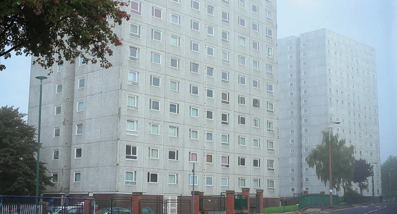 Weekend (2011) movie still: establishing — Two identical high-rise concrete apartment blocks stand side by side under a grey sky. The buildings…