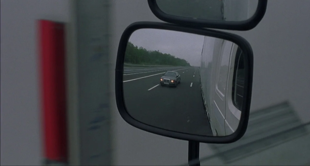 Fat Girl (2001) movie still: closeup — A gray sedan travels on an empty multi-lane highway during the day. The view is captured through a side-view…